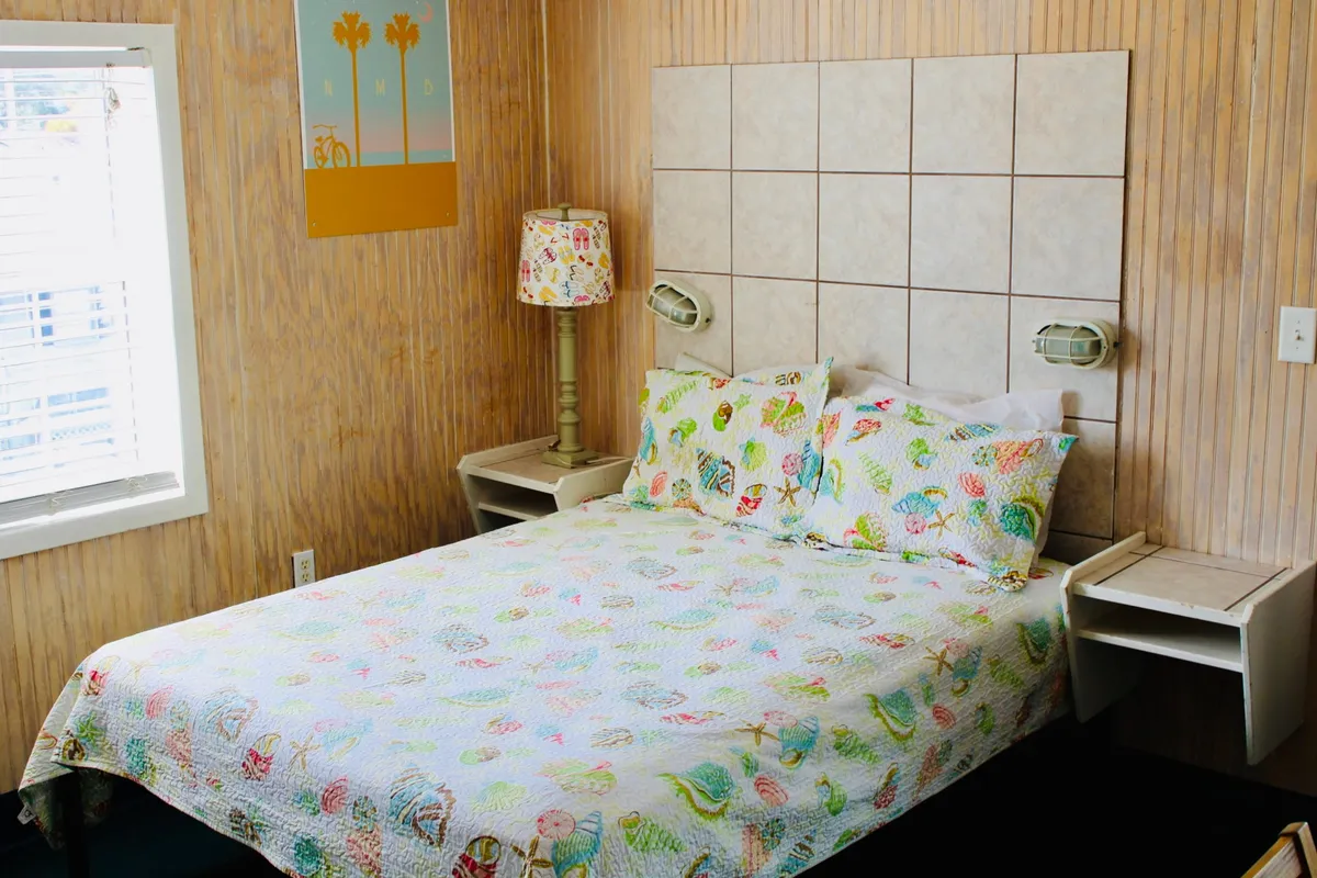 A cheerful, sunlit bedroom area featuring a bed with a colorful quilt, a bedside lamp adorned with a floral pattern, and wall art showcasing palm trees. The space feels inviting and relaxing—perfect for unwinding after a day on the coast.