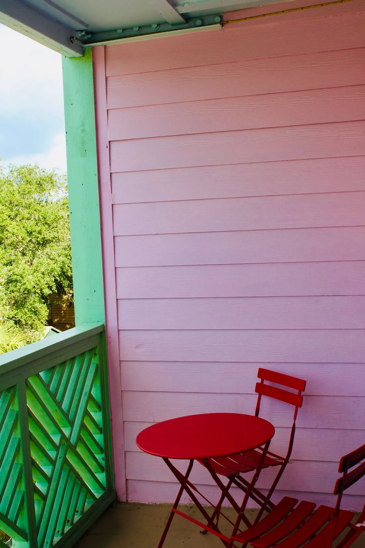 A vibrant exterior seating area with a red table and chairs set against a pink wall and green railing. The scene suggests a spot for enjoying morning coffee, reading, or simply soaking in the fresh air—spaces where guests can relax outdoors with their pets just steps from the apartment.
