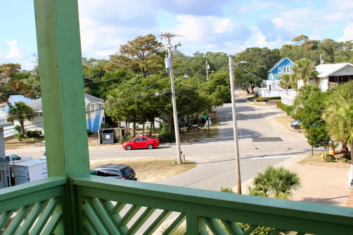 This image presents a relaxed neighborhood scene featuring a blue house, green balcony railing, palm trees, and a red car parked nearby. The foreground captures a sense of casual movement with people walking and exploring the area, suggesting easy access to local attractions and the outdoors. The presence of palm trees and bright daylight conveys a breezy, beach-adjacent atmosphere.
